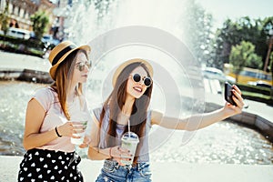 Two young girls in sunglasses and hats near city fountain. Best friends drink cocktails doing self photo and laughing in city stre