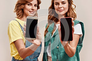 Two young girls, twin sisters in casual wear holding smartphone with blank screen, advertising app  over light