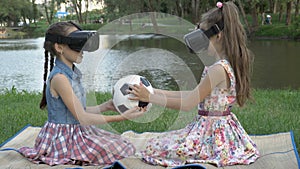 Two young girls in virtual reality glasses together twist and study a soccer ball by touch, sitting in a park against