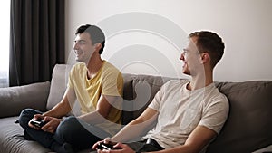 Two young guys playing video games at home, holding joysticks and sitting on the grey sofa in loft interior room.Smiling