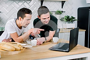 Two young handsome men having breakfast and drinking coffee while using laptop in the kitchen at the morning. Smiling