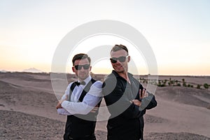 Two young handsome men in shirts and sunglasses standing back to back among the desert, sunset on the background