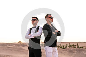 Two young handsome men in shirts and sunglasses standing back to back among the desert, sunset on the background