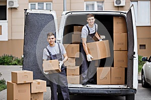 Two young handsome smiling movers wearing uniforms are unloading the van full of boxes. House move, mover service