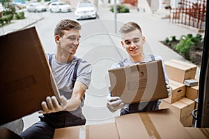 Two young handsome smiling movers wearing uniforms are unloading the van full of boxes. House move, mover service