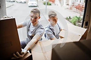 Two young handsome smiling movers wearing uniforms are unloading the van full of boxes. House move, mover service