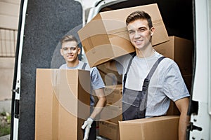 Two young handsome smiling workers wearing uniforms are standing in front of the van full of boxes holding boxes in