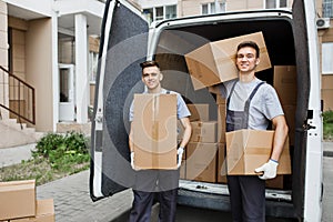 Two young handsome smiling workers wearing uniforms are standing in front of the van full of boxes holding boxes in