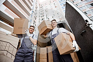 Two young handsome smiling workers wearing uniforms are standing in front of the van full of boxes holding boxes in