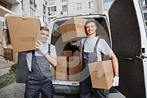 Two young handsome smiling workers wearing uniforms are standing in front of the van full of boxes holding boxes in