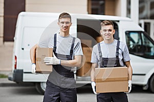 Two young handsome smiling workers wearing uniforms are standing in front of the van full of boxes holding boxes in