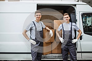 Two young handsome smiling workers wearing uniforms are standing next to the van full of boxes. House move, mover