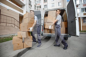 Two young handsome smiling workers wearing uniforms are unloading the van full of boxes. The block of flats is in the
