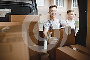 Two young handsome workers wearing uniforms are standing next to the van full of boxes. House move, mover service