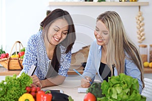 Two young happy women making online shopping for making menu by tablet computer. Friends cooking in the kitchen