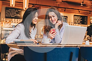 Two young happy women are sitting in cafe at table in front of laptop, using smartphone and laughing.