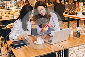 Two young happy women are sitting in cafe at table in front of laptop, using smartphone and laughing.