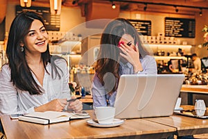 Two young happy women are sitting in cafe at table, using laptop and laughing. On table paper notebook