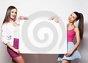 Two young hipster girl friends standing together and holding a blank billboard . Looking at camera