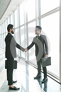 Two young indian businessmen are shaking hands with each other standing against panoramic windows