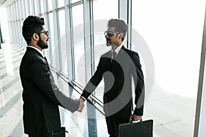 Two young indian businessmen are shaking hands with each other standing against panoramic windows