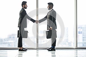 Two young indian businessmen are shaking hands with each other standing against panoramic windows