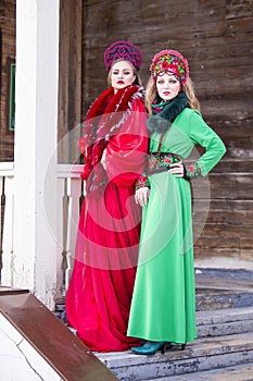 Two Young Ladies in Fashionable Unique Dresses With Kokoshnik Posing Against Old Wooden House in Winter Outdoors