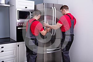 Two Young Male Movers Placing Steel Refrigerator In Kitchen