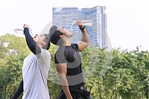 Two young man thirsty drinking water from bottle in their hands after jogging while standing in city park. Handsome male