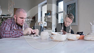 Two young men with disabilities are sculpting clay plates in a home workshop sitting at a large table