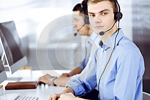 Two young men in headset, sitting at the desk in the modern office, listening to the clients. Call center operators at