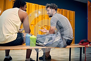 Two young men sitting on bench in dressing room, having a fun conversation after gym workout