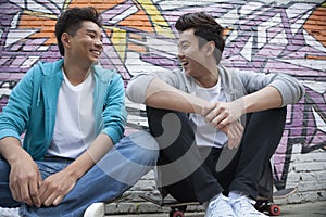 Two young men sitting on their skateboards and hanging out in front of a wall with graffiti