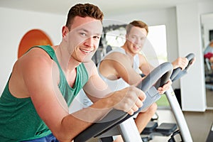 Two Young Men Training In Gym On Cycling Machines Together