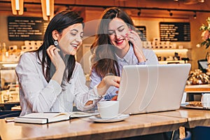 Two young merry businesswomen in shirts sitting in cafe at table and using laptop. Girl is pointing at computer screen