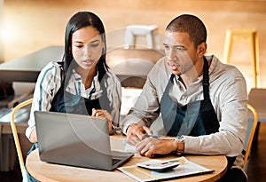 Two young mixed race employees wearing aprons sitting at a table in a coffee shop using a laptop and calculator at work