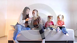 Two young mothers sit with two little daughters on sofa in living room.