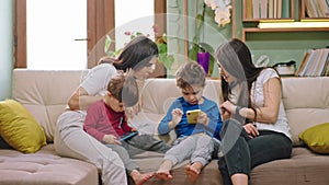 Two young mothers staying at home with their kids they are chatting while sitting on the sofa kids using the smartphone