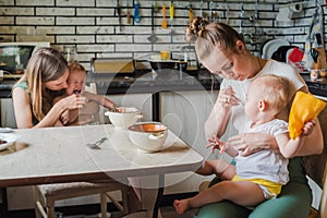 Two young mothers together feed their happy babies milk porridge in the kitchen