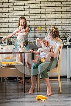 Two young mothers together feed their happy babies milk porridge in the kitchen