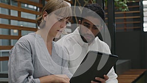 Two young multiracial adults in businesswear talking while using digital tablet in office