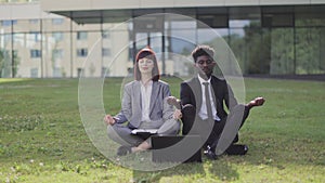 Two young mutli-cultural employees in a lotus pose are sitting on green grass and meditate outside