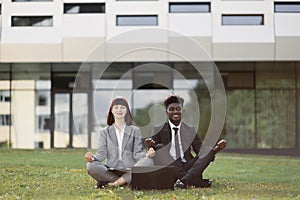 Two young mutli-cultural employees in a lotus pose are sitting on green grass and meditate outside