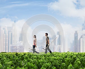 Two young people carrying gardening equipment walking across a green field with plants, cityscape in the background