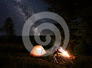 Two young people sit on log near burning fire near tent under powerful fir-tree above the starry sky