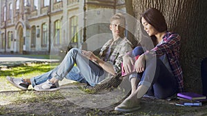 Two young people sitting under tree, girl looking down and guy looking at her