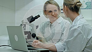 Two young positive scientists working in laboratory using microscope and laptop