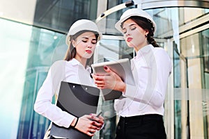 Two young pretty business women industrial engineers in construction helmets with a tablet in hands on a glass building