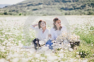 Two young pretty women having picnic with tea in chamomile field