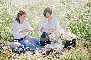 Two young pretty women having picnic with tea in chamomile field